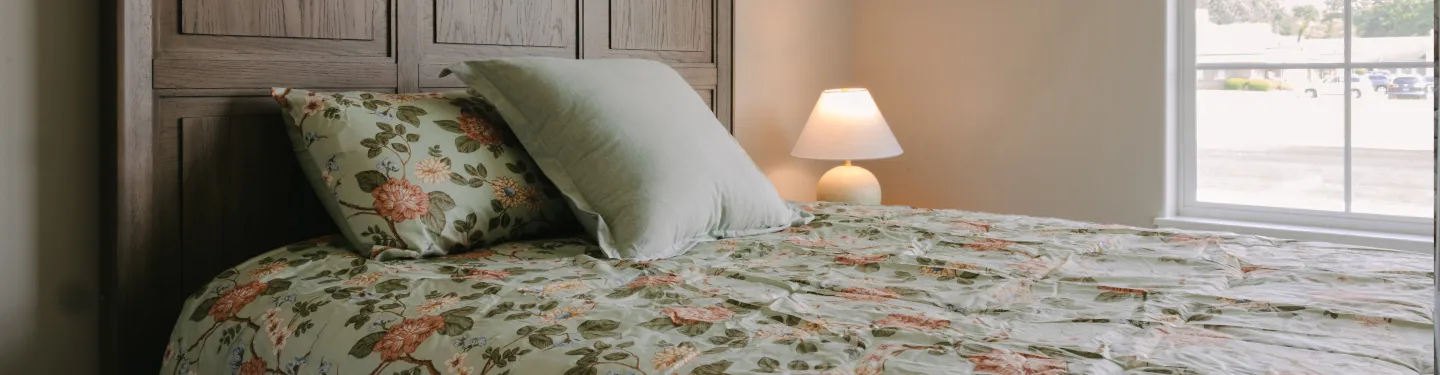 A cozy bed with a floral-patterned bedspread and two pillows is next to a wooden headboard. A small lamp on a nightstand is turned on, and sunlight streams in through a window beside the bed.