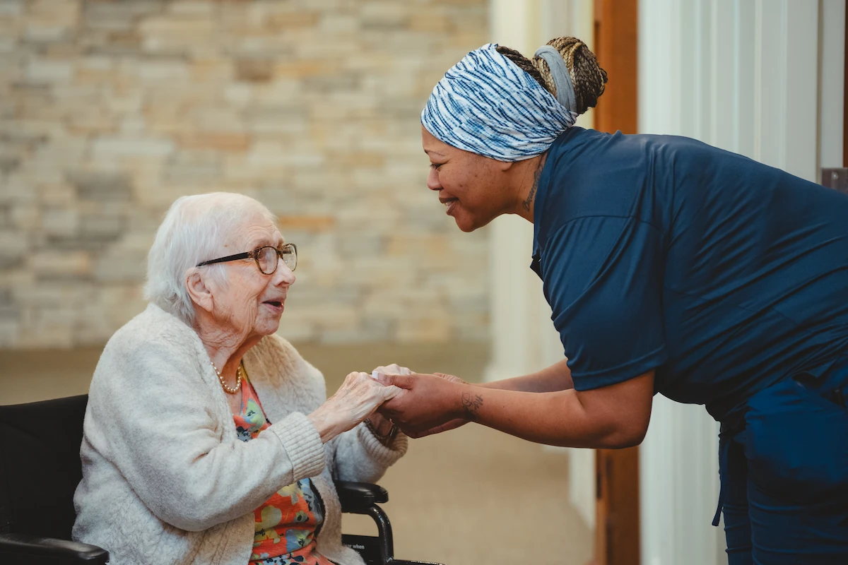 A caregiver smiles warmly and holds hands with an elderly woman in a wheelchair, creating a comforting and supportive atmosphere indoors.
